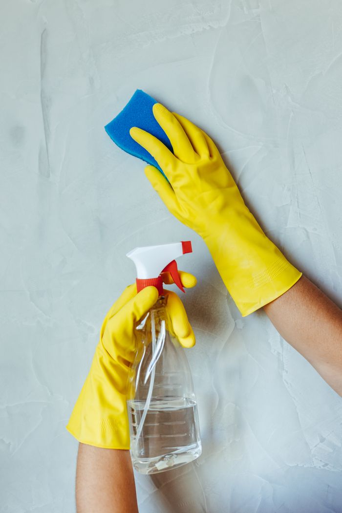 Des mains gantées de jaune nettoient un mur avec une éponge et un vaporisateur.
